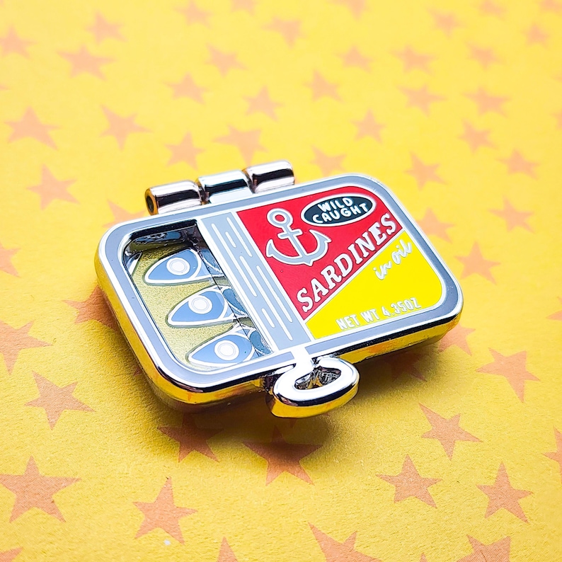 Puede incluir: Pin esmaltado con forma de lata de sardinas con borde plateado. El pin presenta una etiqueta roja y amarilla con las palabras "Wild Caught Sardines en oil" y un ancla. El pin contiene tres sardinas azules y plateadas. Peso neto 123g.