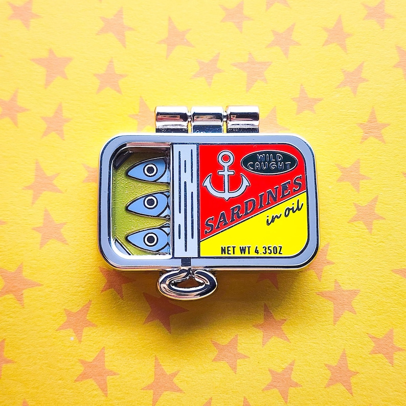Puede incluir: Un pin de lata de sardinas plateada con una etiqueta roja y amarilla que dice "Sardines in oil" y "Wild Caught". La lata contiene tres peces de dibujos animados con ojos azules sobre un fondo amarillo. Peso neto: 123 gramos.