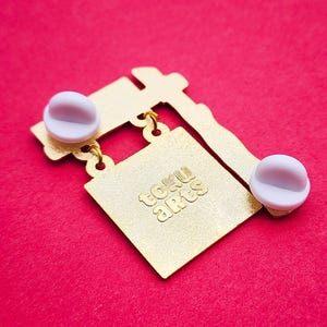 May include: Gold-colored metal pin with a square design and the text "toku arts" in a stylized font. The pin has two white pin backs and a red background.