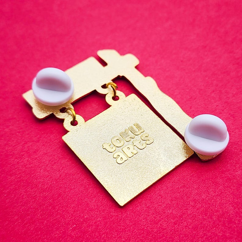 May include: Gold-colored metal pin with a square design and the text "toku arts" in a stylized font. The pin has two white pin backs and a red background.