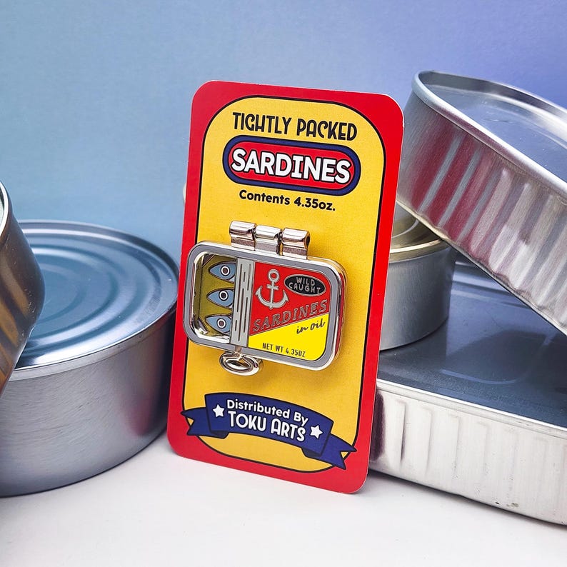 Puede incluir: Un pin con tem&aacute;tica de sardinas en una tarjeta de respaldo roja y amarilla. El pin es una lata de sardinas en miniatura con las palabras "Sardines in oil" y "Wild Caught". La tarjeta dice "Tightly Packed Sardines" y "Contenido 123g".