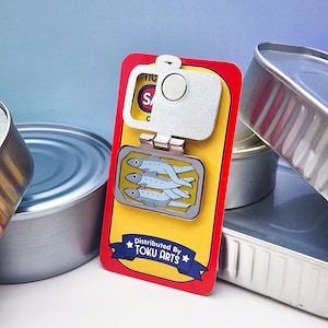 Puede incluir: Un pin con forma de lata de sardinas con un abridor de botellas plateado. El pin presenta un fondo amarillo con una imagen rectangular de sardinas. El texto "Distributed By TOKU ARTS" est&aacute; en una pancarta azul.