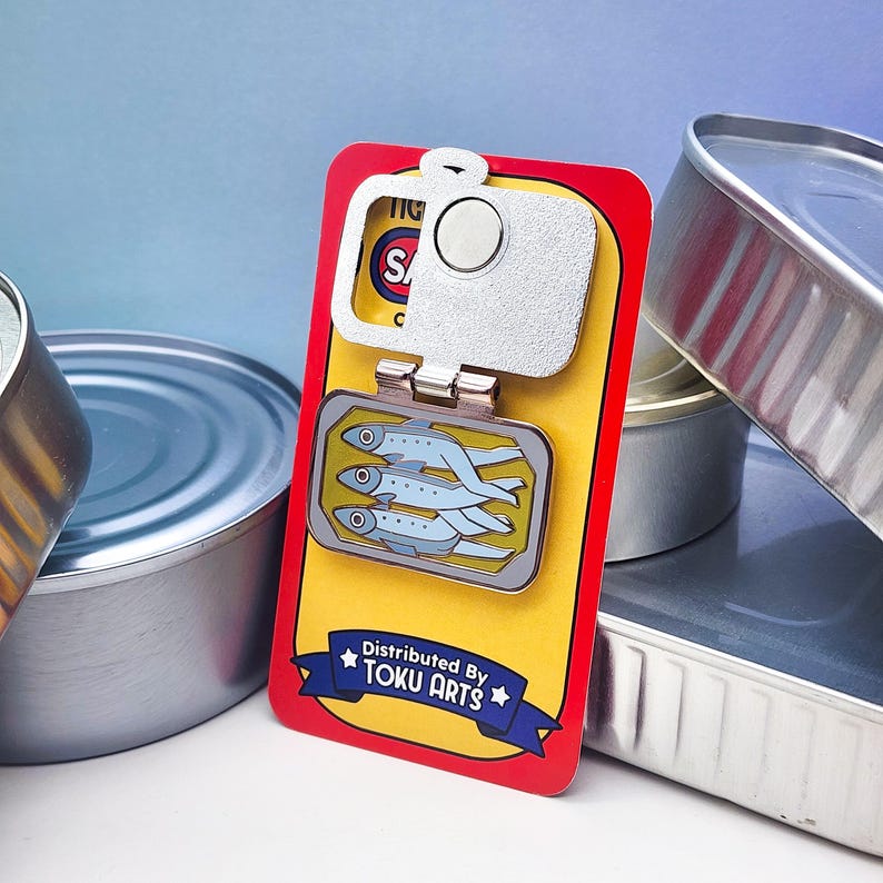 Puede incluir: Un pin con forma de lata de sardinas con un abridor de botellas plateado. El pin presenta un fondo amarillo con una imagen rectangular de sardinas. El texto "Distributed By TOKU ARTS" est&aacute; en una pancarta azul.