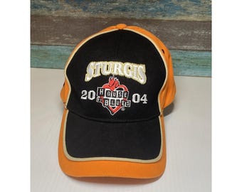 May include: Orange and black baseball cap with the text "Sturgis 2004 House of Blues". The cap has a curved brim and a structured crown. The design includes a heart-shaped logo with flames.