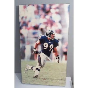 May include: A framed print featuring an American football player in a navy blue uniform with the number 99. The player is running on a green field, with a blurred crowd in the background. The print is rectangular and vertical.