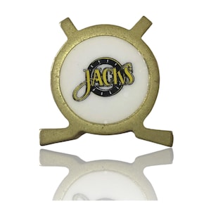 May include: A gold-coloured metal pin with a white centre featuring the word "JACKS" in yellow script. The pin has a circular design with four protruding tabs. The text is surrounded by a black and yellow design.