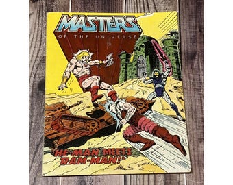 Puede incluir: Portada de cómic vintage de Masters of the Universe. La portada presenta una escena de acción dinámica con He-Man, Ram-Man y Skeletor. El título está en fuente azul y blanca sobre un fondo amarillo. El cómic está sobre una superficie de madera.