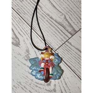 Può includere: Una collana con pendente stravagante con una statuina di un personaggio in bicicletta rossa. Il personaggio indossa un vestito rosa e un cappello viola. Il pendente è attaccato a un cordino nero e posto su uno sfondo blu a forma di nuvola.