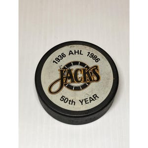 May include: A black hockey puck with a white and black circular design. The design includes the text "1936 AHL 1986" and "Jacks" in a stylized font. The text "50th YEAR" is also present.