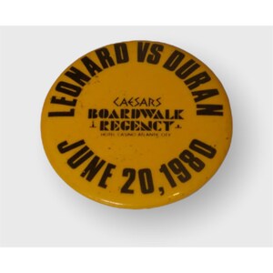 May include: A round yellow pinback button featuring black text that reads LEONARD VS DURAN, CAESARS BOARDWALK REGENCY HOTEL CASINO ATLANTIC CITY, and JUNE 20, 1980. The button has a smooth, glossy surface and a circular shape with bold lettering.