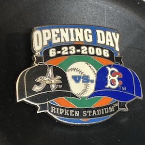 May include: A baseball pin with the text "Opening Day" and the date "6-23-2006". The pin shows baseball caps for the Atlanta Braves and Boston Red Sox, with a baseball in the middle. The words "VS." and "Ripken Stadium" are also visible.