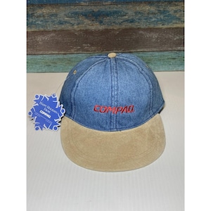 May include: A blue denim baseball cap with a tan suede brim. The cap has the word "COMPAQ" embroidered in red on the front.