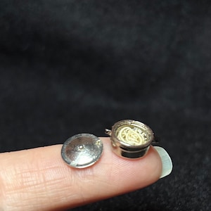May include: A miniature silver pot with a lid, filled with white noodles. The pot is sitting on a finger.