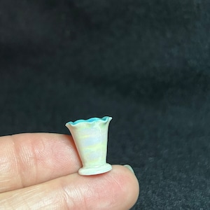 May include: A small, white ceramic vase with a blue rim and yellow and purple stripes. The vase has a scalloped top and a round base.