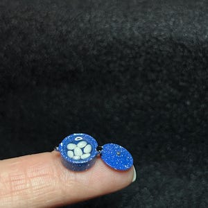 May include: A small blue and white ceramic container with a lid. The container is filled with white beads and the lid is open. The container is about the size of a fingernail.