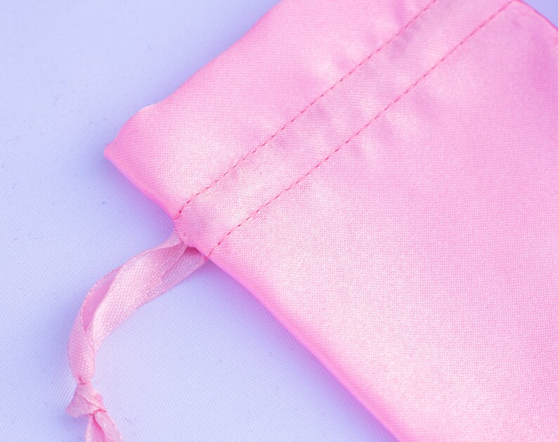 May include: A pink satin drawstring bag with a white cord. The bag is made of soft, silky fabric and has a smooth, shiny finish.