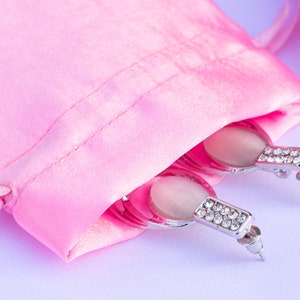 May include: A pair of silver earrings with white oval stones and clear crystal accents are displayed in a pink satin pouch.