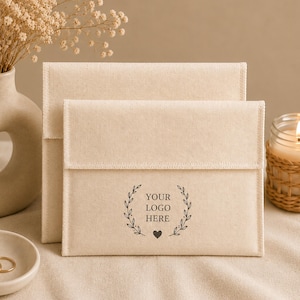 May include: Two rectangular beige fabric envelopes with a flap closure. The front envelope features a black printed design consisting of two curved leafy branches framing the text "YOUR LOGO HERE" with a small heart icon centered below the text.