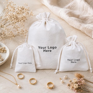 May include: Three off-white cotton drawstring pouches of varying sizes, each featuring the black printed text Your Logo Here. The pouches are arranged with gold-toned jewelry, including a necklace, two rings, and a pair of pearl earrings.