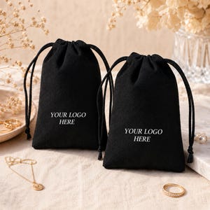 May include: Two black velvet drawstring pouches featuring the white text YOUR LOGO HERE printed on the front. Each pouch has black cord ties at the top, providing a secure closure for small items like jewelry or accessories.