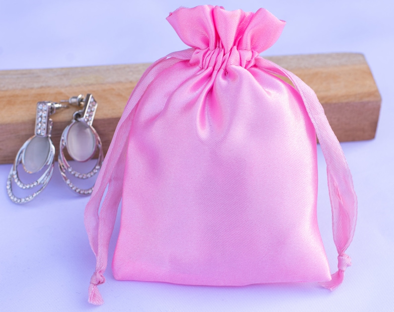 May include: A pink satin drawstring bag with a white background. The bag is empty and has a small opening at the top.