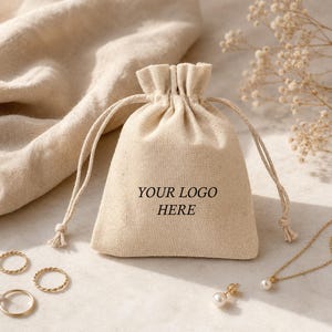 Può includere: Un sacchetto beige con coulisse con la scritta nera "YOUR LOGO HERE". Gioielli dorati, tra cui anelli, una collana e orecchini con perle, sono disposti attorno al sacchetto. Lo sfondo presenta un tessuto beige e fiori secchi.