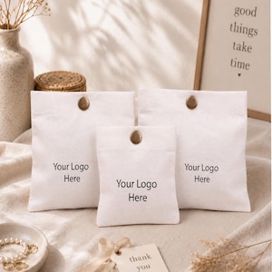 May include: Three off-white fabric pouches of varying sizes, each featuring a single metallic snap button closure at the top centre and the black printed text Your Logo Here centred on the front of each individual pouch.