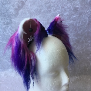 May include: A pair of faux fur cat ears in shades of purple, pink, and white, attached to a black headband. The ears have a fluffy texture and a silver charm with a moon and star design. The ears are displayed on a white foam head.