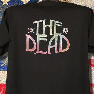 May include: Black t-shirt with a rainbow gradient print of "THE DEAD" in a stylized font. The print includes a skull and crossbones and a rose.