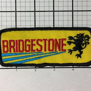 Könnte beinhalten: Ein gelber gestickter Aufnäher mit dem Text "BRIDGESTONE" in Rot und einem schwarzen Löwenlogo. Der Aufnäher hat einen schwarzen Rand und drei blaue Streifen.