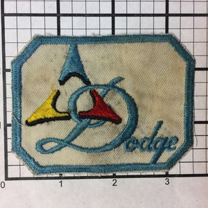 Puede incluir: Un parche bordado azul, rojo y amarillo con la palabra "Dodge" en letra cursiva azul.