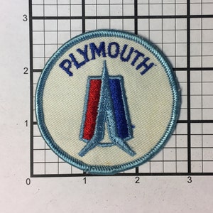 Può includere: Un cerotto circolare bianco con un bordo blu. Al centro del cerotto è presente una freccia stilizzata blu e rossa con la parola "PLYMOUTH" in lettere blu attorno alla freccia.