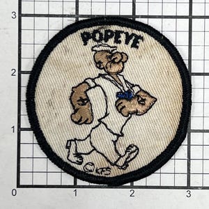 Op de afbeelding: Een witte stoffen patch met zwarte stiksels en een zwarte rand. De patch heeft een cartoon afbeelding van Popeye, een zeeman met een wit shirt, een blauwe broek en een rode pet. De tekst "POPEYE" is in zwart boven de afbeelding gedrukt.