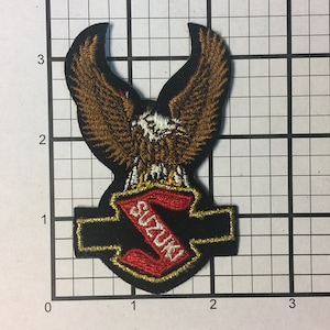 May include: A black and gold embroidered patch with a red "Suzuki" logo and a brown eagle with white accents.
