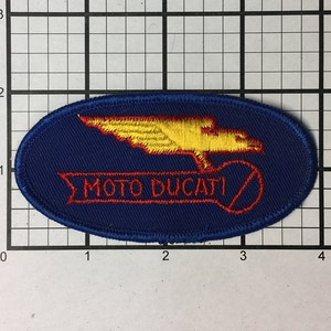 Może przedstawiać: Naszywka w kształcie owalu, w kolorze niebieskim, z haftowanym żółtym logo skrzydła i napisem "MOTO DUCATI" w kolorze czerwonym.
