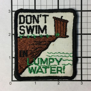 Może przedstawiać: Biała naszywka haftowana z czarną lamówką. Naszywka przedstawia brązowy szalet z zielonym dachem i tekstem "DON'T SWIM IN LUMPY WATER!" w kolorze zielonym i czarnym.