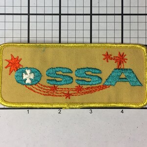 May include: A yellow embroidered patch with the text "OSSA" in teal blue lettering. The patch features a stylized firework design with orange and red accents.