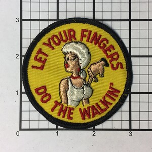 May include: A yellow embroidered patch with a black border. The patch features a cartoon woman with a white hat and a red dress. The text on the patch reads "Let Your Fingers Do The Walkin".