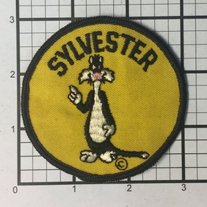 May include: A yellow circular patch with black stitching around the edges. The patch features a cartoon image of Sylvester the cat from Looney Tunes. The cat is white with black markings and is standing on his hind legs with his front paw raised. The word "SYLVESTER" is written in black around the cat's head.