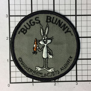 May include: A round, gray fabric patch with black stitching. It features a cartoon image of Bugs Bunny holding a carrot and the text "Bugs Bunny" in a black, cursive font. The text "Warner Bros. Inc. 1973 Klandex" is also visible in black.