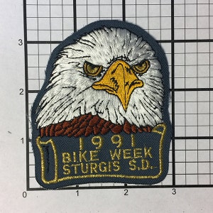 May include: A blue embroidered patch featuring a white bald eagle head with a yellow beak. The patch has the text "1991 BIKE WEEK STURGIS S.D." in gold thread.