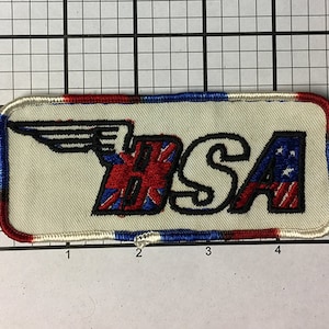 May include: A white embroidered patch with a winged logo and the text "USA". The logo is a stylized "U" with a Union Jack flag design and an "S" with a US flag design. The patch has a red, white, and blue border.