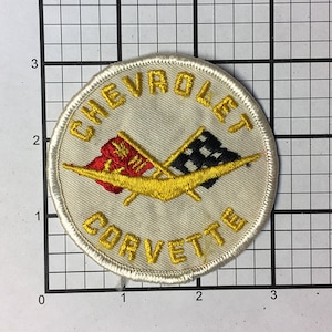May include: A white embroidered patch with a yellow Corvette logo and the words "Chevrolet Corvette" in yellow lettering. The logo features two crossed flags, one red and white and the other black and white.