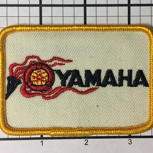 May include: A yellow embroidered patch with the word "YAMAHA" in black lettering. The logo features a red and yellow stylized flame design.