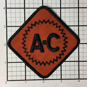 Puede incluir: Un parche de color marrón en forma de diamante con un borde negro y un diseño de estrella negra y blanca. Las letras "A-C" están bordadas en negro en el centro del parche.