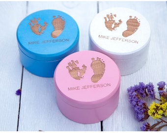 May include: Three round wooden keepsake boxes in blue, white, and pink. Each lid features a natural wood-toned engraved handprint and footprint design with the text MIKE JEFFERSON centered below the prints in a clean, sans-serif font style.