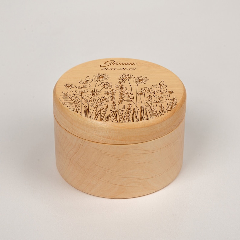 Op de afbeelding: Een kleine, ronde houten doos met een deksel. Het deksel is gegraveerd met een bloemenmotief en de tekst "Jenna 2011-2019".
