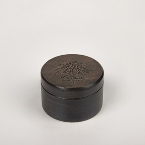 Op de afbeelding: Een ronde houten doos met een donkerbruine vlek. Het deksel heeft een gegraveerd beeld van een cannabisplant en de tekst "James Bost" met een klein logo eronder.