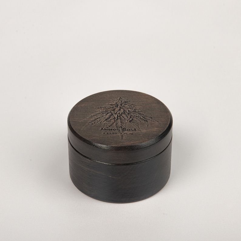Op de afbeelding: Een ronde houten doos met een donkerbruine vlek. Het deksel heeft een gegraveerd beeld van een cannabisplant en de tekst "James Bost" met een klein logo eronder.