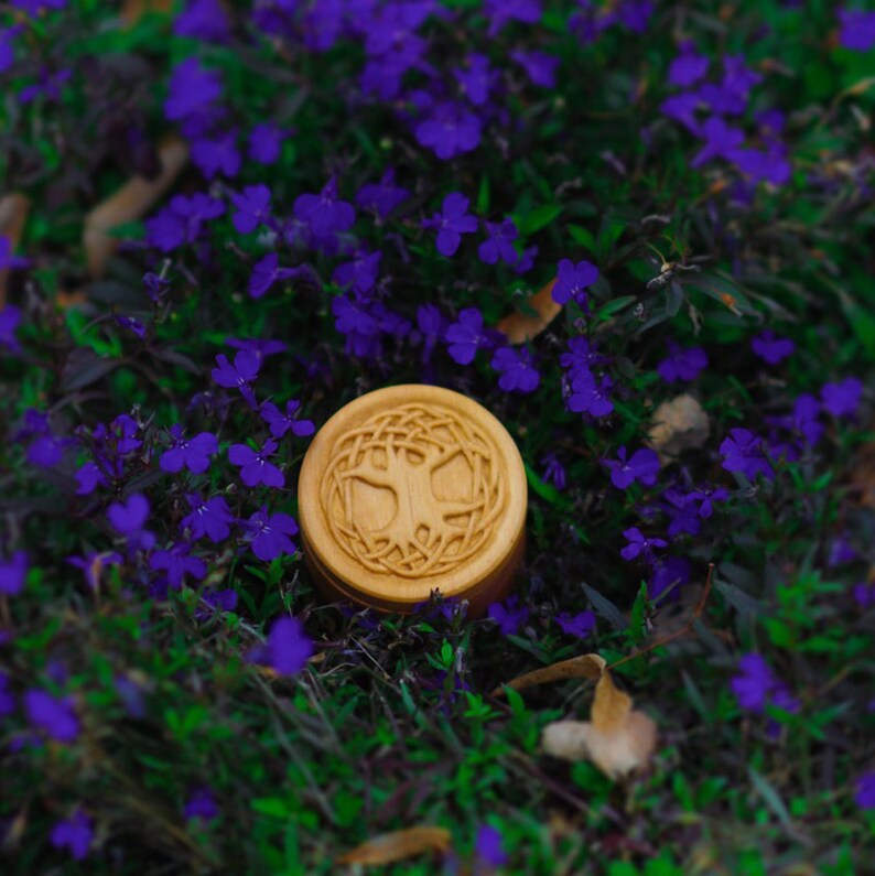 Pu&ograve; includere: Una scatola di legno con un design ad albero della vita intagliato, circondata da fiori viola.